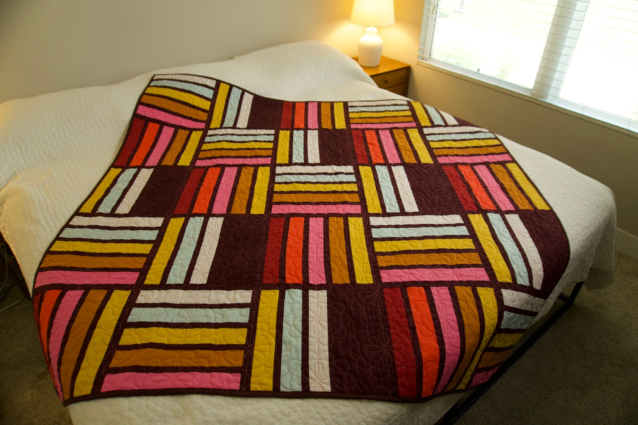 A vibrant striped patchwork quilt in mustard, coral, and blush laid across a bed in a sunlit bedroom